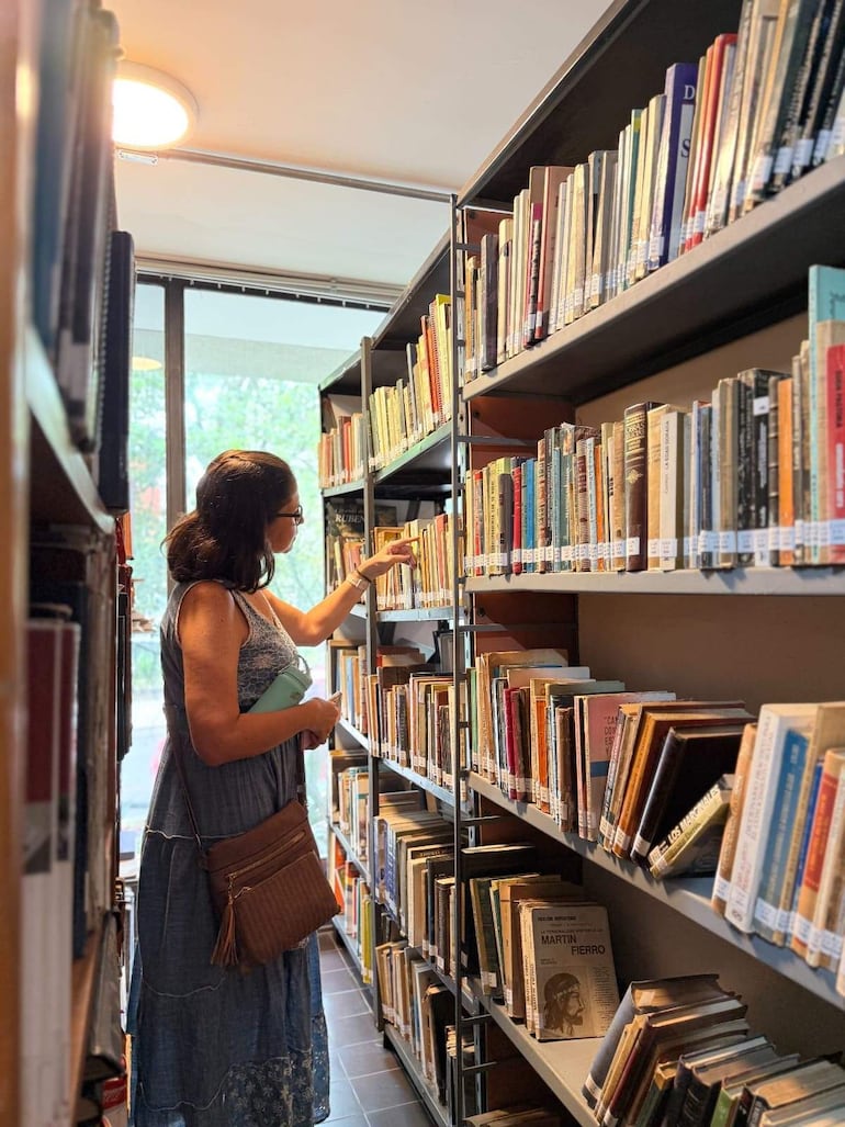 Mujer con vestido azul claro examinando un libro en una biblioteca con estanterías metálicas y luz natural.