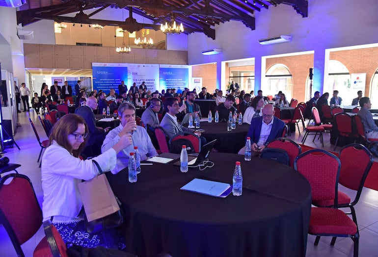 Mujer con cabello largo y gafas sentada, manipulando una bolsa, mientras hombres interactúan en un salón bien iluminado.