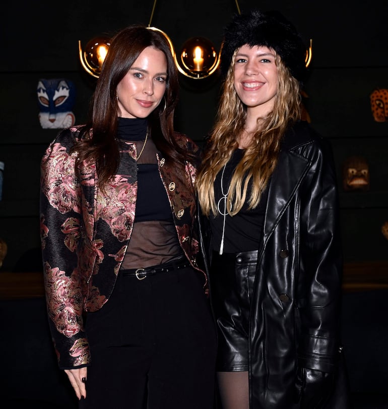 Dos mujeres sonrientes, una con chaqueta floral y blusa negra, otra con abrigo de cuero y gorro negro, en ambiente decorativo.