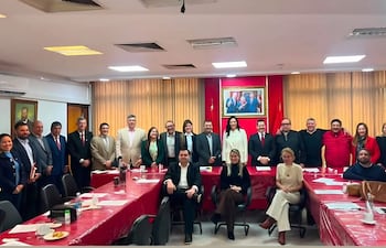 Carmen Dejesús Giménez de Ovando y otros asistentes en trajes formales, en un salón con paredes rojas, discuten documentos.