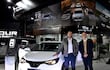 Dos hombres sonríen junto a un vehículo eléctrico blanco, uno con chaqueta gris y camisa blanca, el otro con suéter beige y chaqueta oscura.