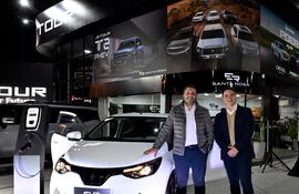 Dos hombres sonríen junto a un vehículo eléctrico blanco, uno con chaqueta gris y camisa blanca, el otro con suéter beige y chaqueta oscura.