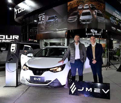 Dos hombres sonríen junto a un vehículo eléctrico blanco, uno con chaqueta gris y camisa blanca, el otro con suéter beige y chaqueta oscura.