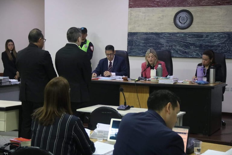 Tres individuos en el tribunal: dos mujeres y un hombre. Una mujer habla, el hombre toma notas y otra observa.