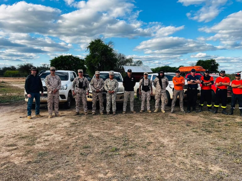Catorce personas en zona rural, algunas con uniformes de camuflaje y otras en camisetas, esperando en un día nublado.