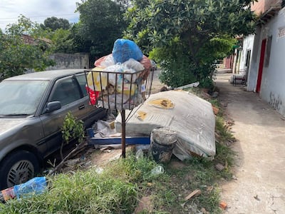 Contenedor de basura desbordante junto a un automóvil gris, casas blancas y un entorno descuidado en el barrio Itay.