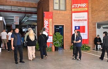 Grupo de personas en el centro de convenciones, una mujer de vestido negro y un hombre con chaleco oscuro observan el ambiente.