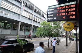 Cartel luminoso en una calle comercial con tasas de cambio de dólar, euro y real, entre árboles y personas en movimiento.