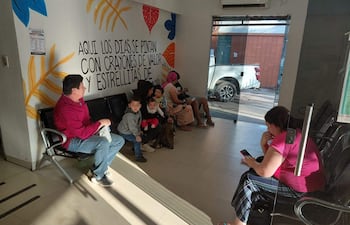 Una mujer sostiene a un bebé mientras tres niños juegan cerca, un hombre relajado a su izquierda en un ambiente colorido de espera.