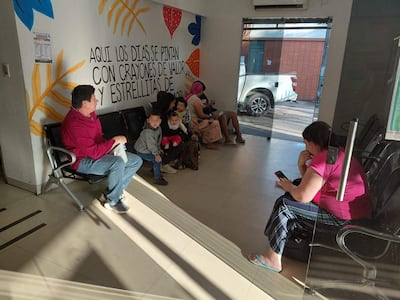 Una mujer sostiene a un bebé mientras tres niños juegan cerca, un hombre relajado a su izquierda en un ambiente colorido de espera.