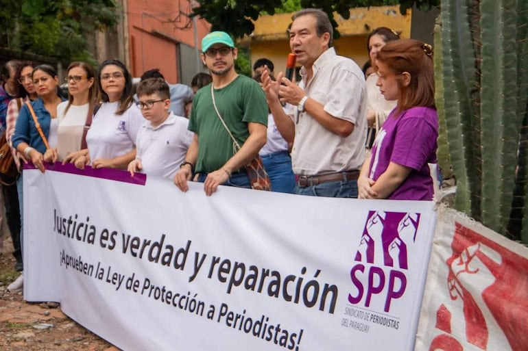 Hombre con camisa clara habla con micrófono, rodeado de asistentes que sostienen un banner sobre protección a periodistas.