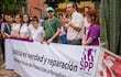 Hombre con camisa clara habla con micrófono, rodeado de asistentes que sostienen un banner sobre protección a periodistas.