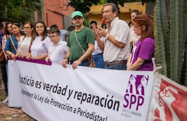 Hombre con camisa clara habla con micrófono, rodeado de asistentes que sostienen un banner sobre protección a periodistas.
