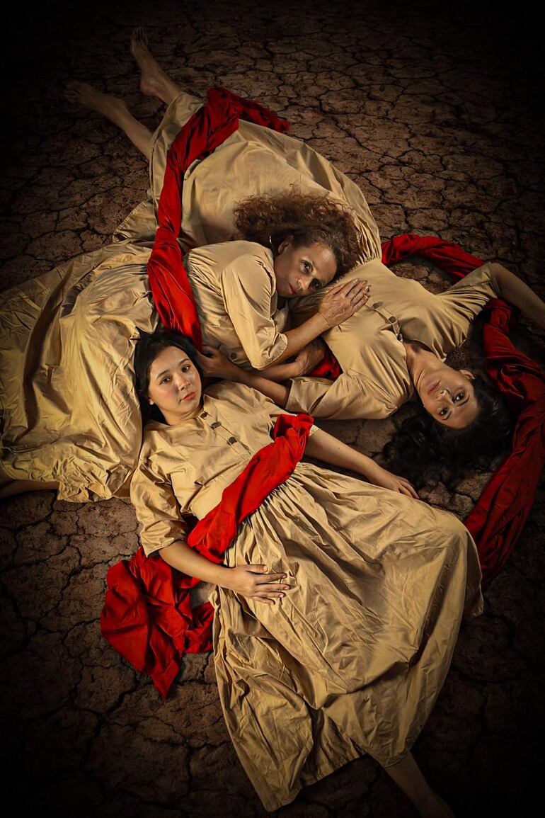 Marcela Gilabert, Eli Marín y Roxana Lescano en vestidos beige y cintas rojas, reclinadas sobre terreno seco, con expresiones serias.
