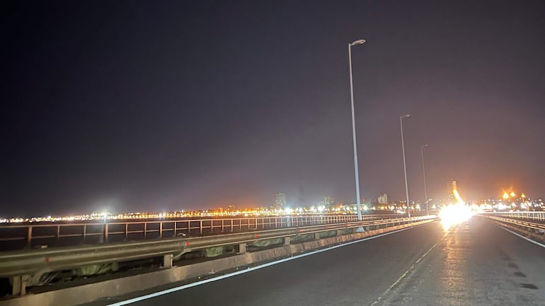 Vista nocturna del puente San Roque iluminado desde un costado, sin personas visibles y con una carretera en primer plano.