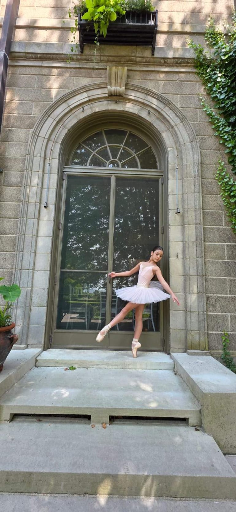 Bailarina en tutú blanco y zapatillas, en pose de danza con fondo de plantas y puerta de cristal en un día soleado.