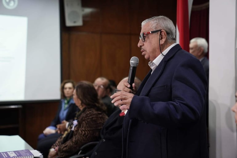 Isaías Fretes, con abrigo oscuro y gafas, habla con micrófono ante un público atento en un auditorio decorado con una bandera.