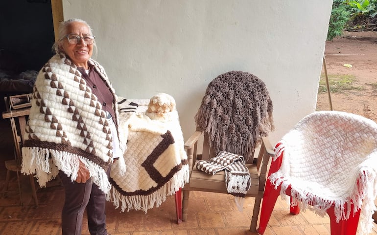 Fermina Fernández sonríe, vestida con blusa oscura y manta tejida, rodeada de artesanías de tejido en un entorno exterior.