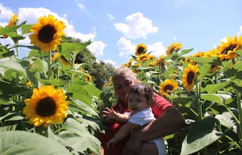 Mujer en camiseta roja sonríe mientras sostiene a su bebé, rodeados de girasoles bajo un cielo despejado.