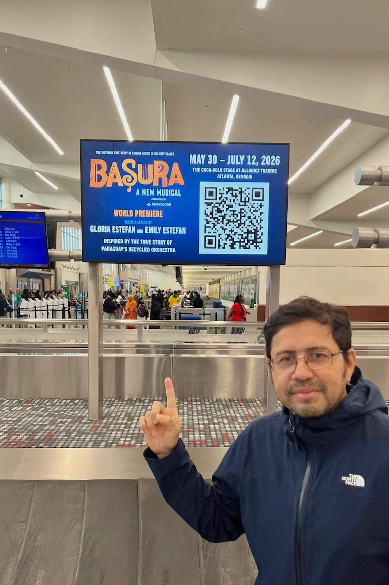 Hombre con chaqueta oscura y gafas señala un cartel que anuncia el musical 'Basura' en un aeropuerto moderno.
