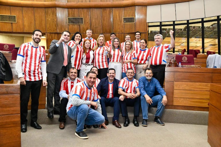 Diputados sonrientes en camisetas rayadas rojas y blancas, posando con brazos levantados en un ambiente festivo dentro del recinto legislativo.