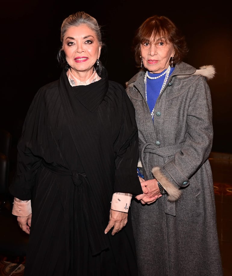 Dos mujeres, una con abrigo gris y blusa azul y otra en elegante atuendo negro, conversan en ambiente de homenaje.