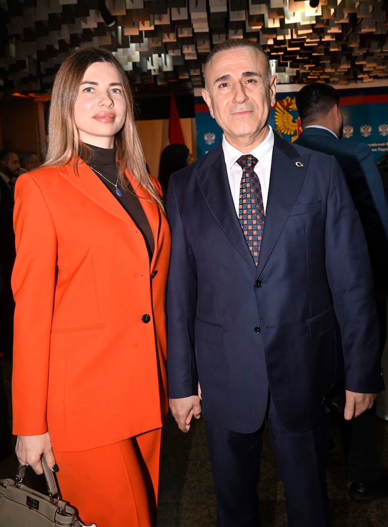 Mujer con traje naranja y bolso, sonriente, junto a hombre en traje azul oscuro y corbata colorida en evento formal.