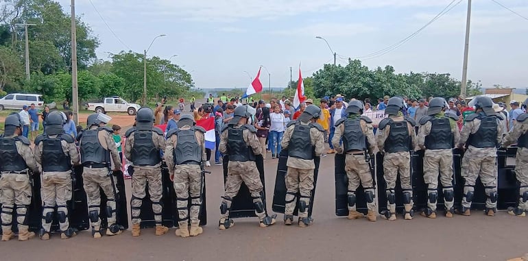 Agentes de seguridad en fila con escudos mientras campesinos con banderas paraguayas se manifiestan.