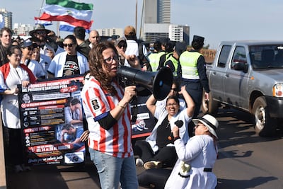 Mujer con camiseta a rayas rojas y blancas sostiene un megáfono, rodeada de manifestantes con carteles y banderas en un día soleado.