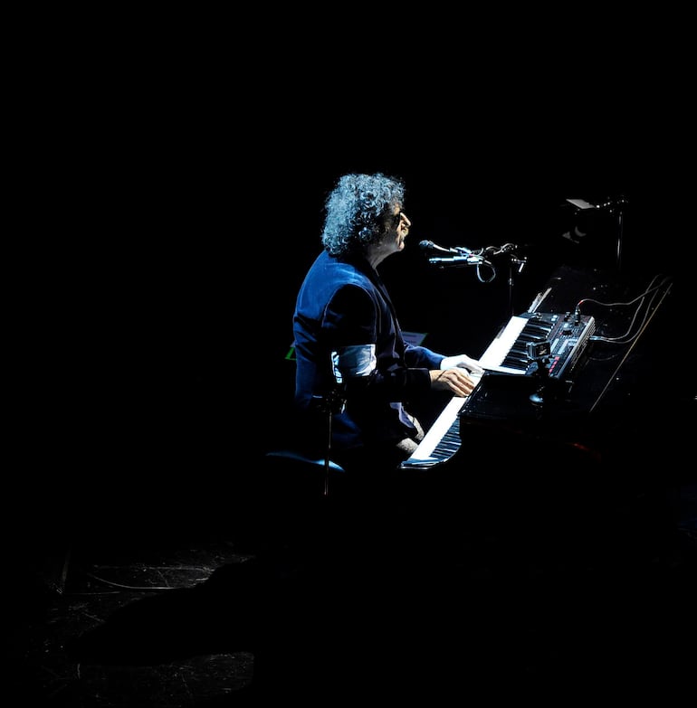 Charly García con cabello rizado y chaqueta oscura, tocando el piano en un ambiente iluminado de manera íntima.