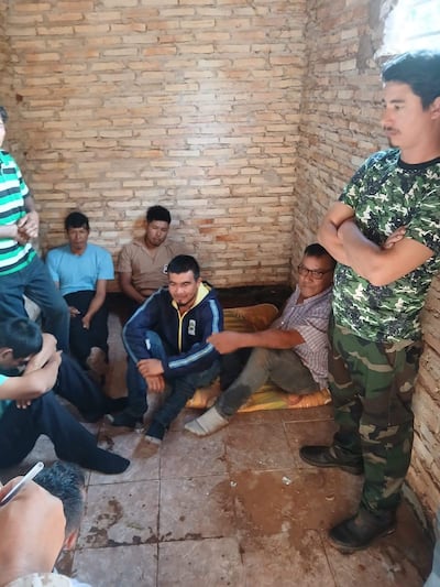 Hombre de pie en camisa de camuflaje habla a otros sentados, algunos atentos y otros relajados, en habitación sencilla.