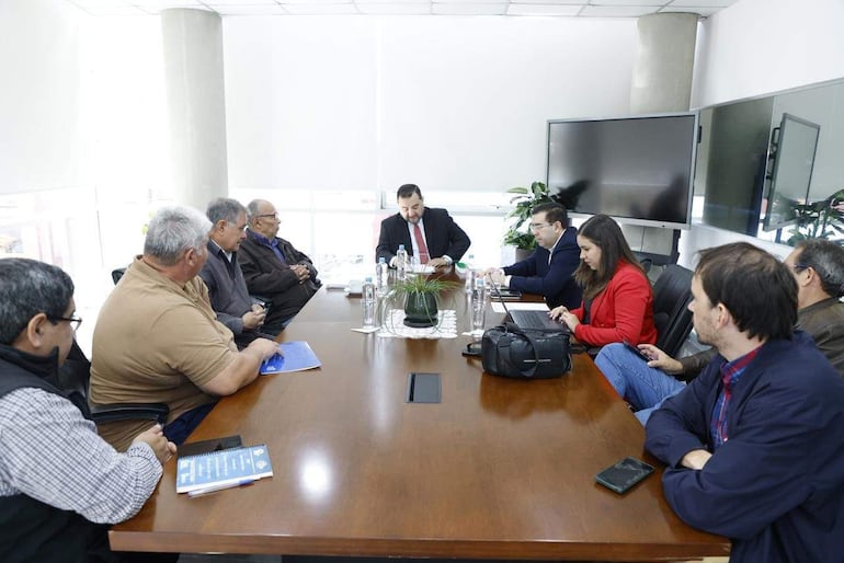 César Segovia y otros en reunión, discutiendo documentos. Asistentes atentos, mujer con laptop a la derecha, ambiente moderno.