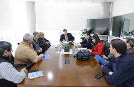 César Segovia y otros en reunión, discutiendo documentos. Asistentes atentos, mujer con laptop a la derecha, ambiente moderno.