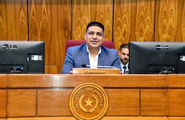 Hugo Meza sonriente, vestido con blazer azul y camisa blanca, sentado en escritorio con placa que dice 'Presidente'.