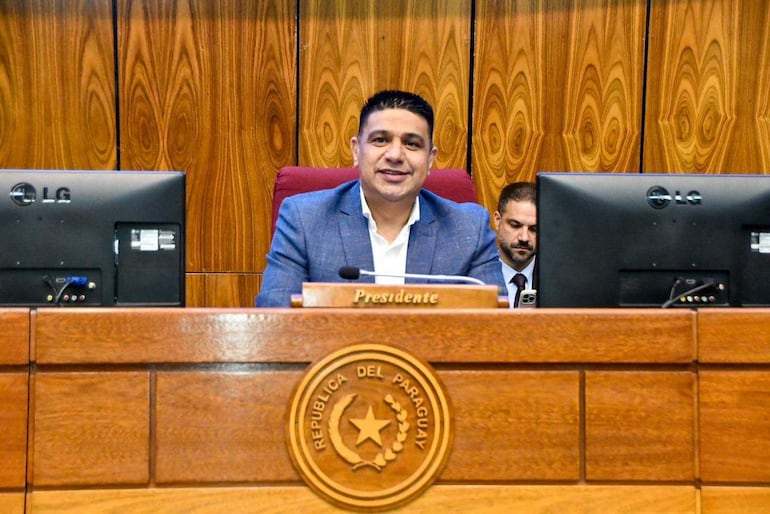 Hugo Meza sonriente, vestido con blazer azul y camisa blanca, sentado en escritorio con placa que dice 'Presidente'.