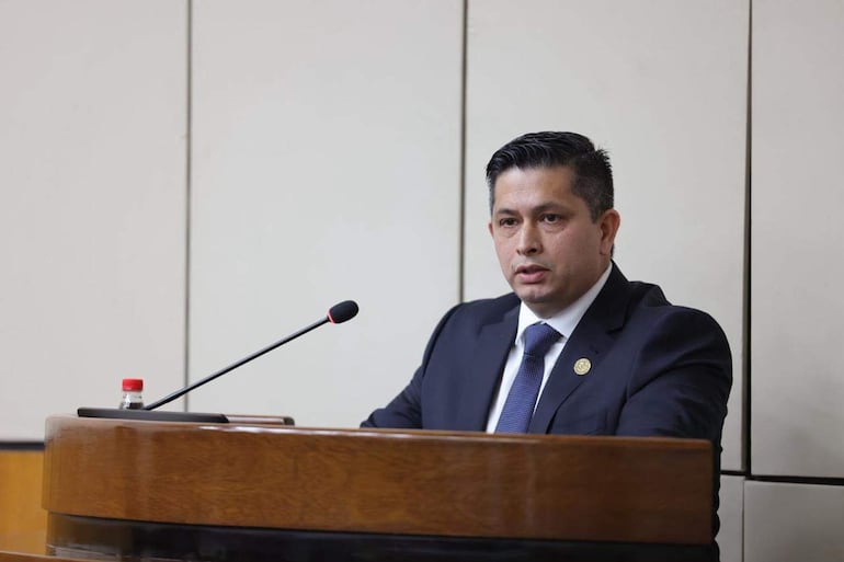 Armindo Torres, vestido con traje oscuro y corbata azul, habla frente a un micrófono en un salón de reuniones formal.