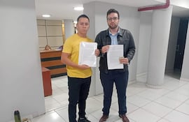 Bruno Martínez con camiseta amarilla y Maximiliano Seiferheld en camisa azul y chaqueta negra, mostrando documentos en oficina austera.