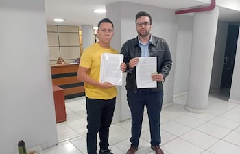 Bruno Martínez con camiseta amarilla y Maximiliano Seiferheld en camisa azul y chaqueta negra, mostrando documentos en oficina austera.