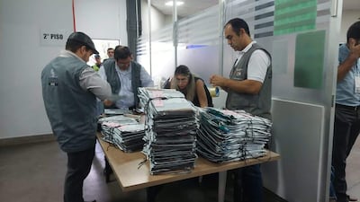 Hombre con gorra organiza documentos, mientras otro revisa papeles. Mujer con gafas observa. Ambiente formal y ordenado.