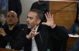 Hombre de barba y chaqueta negra habla gesticulando, mientras más personas escuchan atentamente en un ambiente formal.
