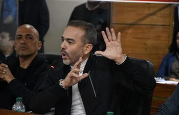 Hombre de barba y chaqueta negra habla gesticulando, mientras más personas escuchan atentamente en un ambiente formal.