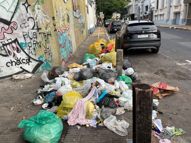 Montículo de basura plástica de colores en la acera, con graffiti en una pared cercana y vehículos estacionados al fondo.