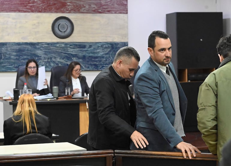 Dos mujeres detrás de un escritorio en el tribunal, mientras tres hombres discuten, uno en traje claro y corbata oscura.