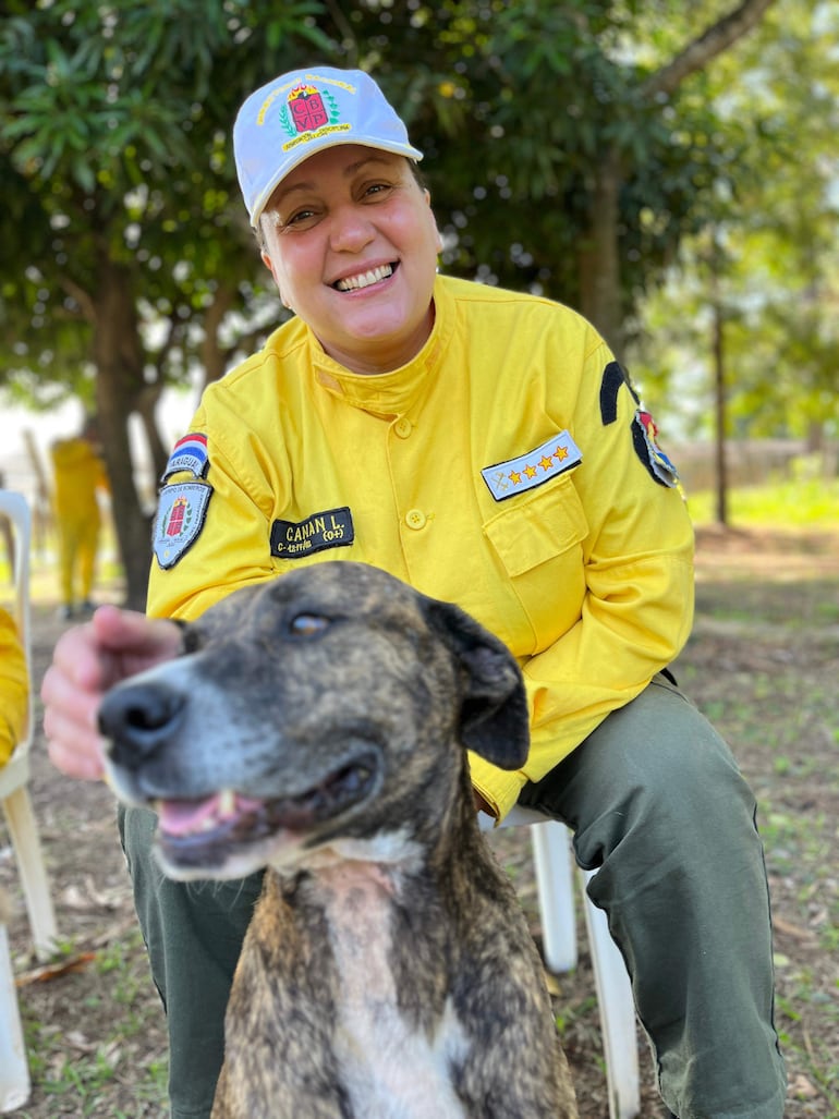 Lorena Canan, bombera sonriente con camisa amarilla y gorra celeste acaricia a su perro marrón en un entorno natural.