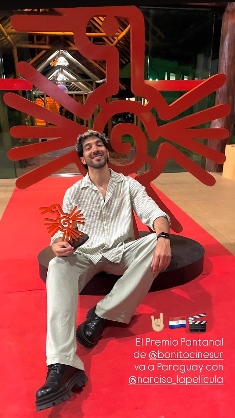 Diro Romero sentado sobre alfombra roja, con trofeo en mano, vistiendo camisa de rayas y pantalones claros, sonriente.