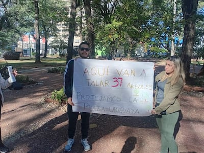 Hombre con gafas de sol y mujer con abrigo sostienen cartel sobre tala de árboles en la Plaza Uruguaya. Tercer observador en el fondo.