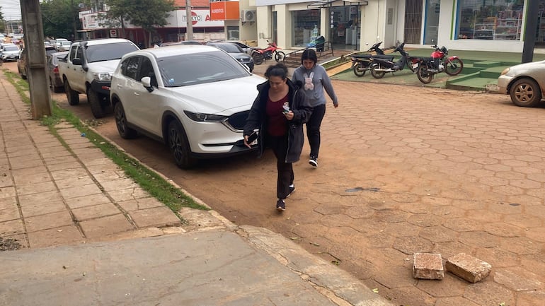 Dos mujeres, una con chaqueta negra y otra con suéter gris, caminan por la acera con expresión triste.