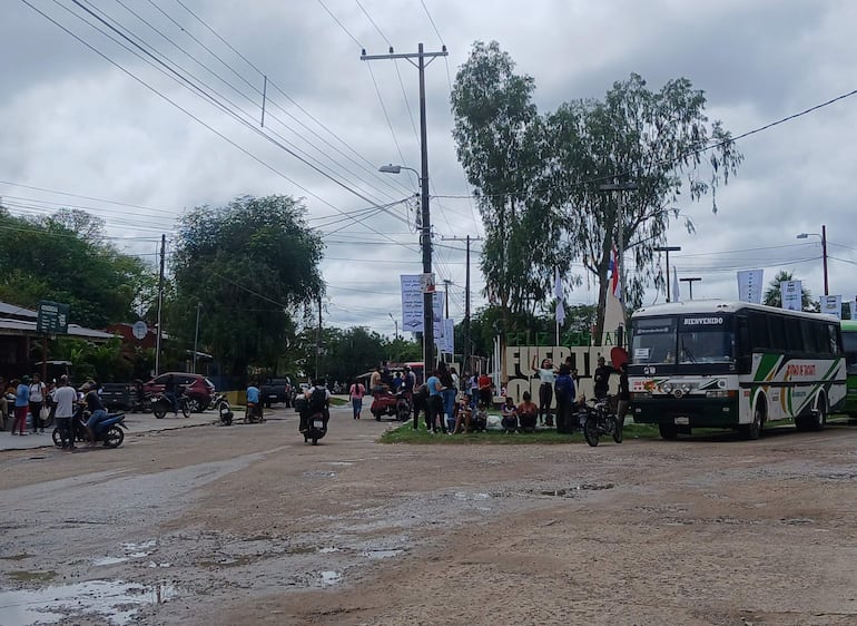 Grupo de personas en la acera, algunas sentadas, con un autobús de línea a la derecha bajo un cielo nublado.