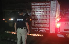 Policía de espaldas frente a camión cubierto, con camiseta negra que dice 'POLICÍA', en ambiente nocturno y camioneta con luces encendidas.