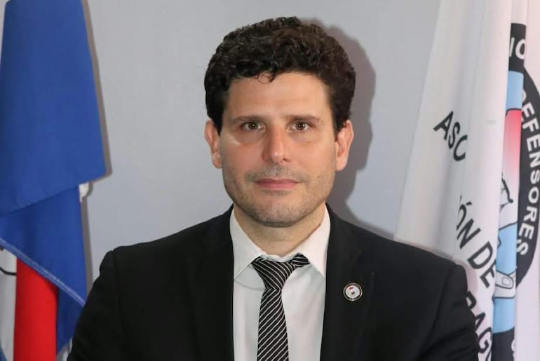 Martín Muñoz con traje negro y corbata blanca, frente a fondo con banderas, en posición formal y tranquila.